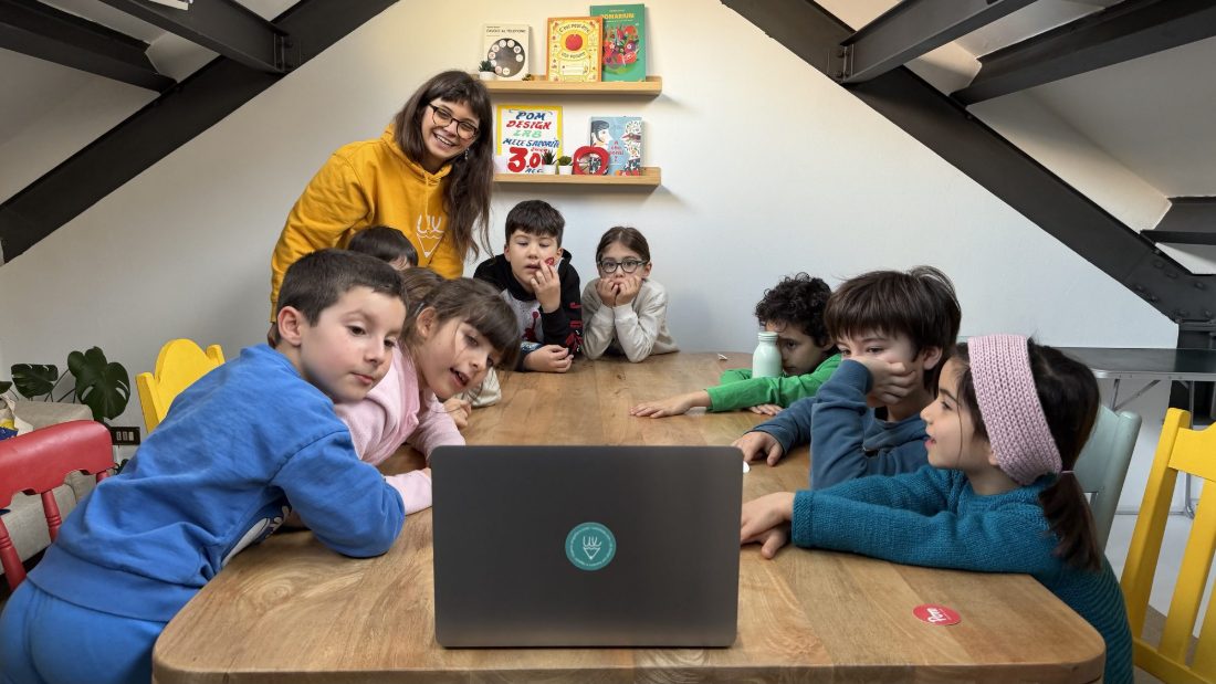 Un gruppo di bambini attorno al tavolo guarda lo schermo di un laptop, con una mentor dietro di loro