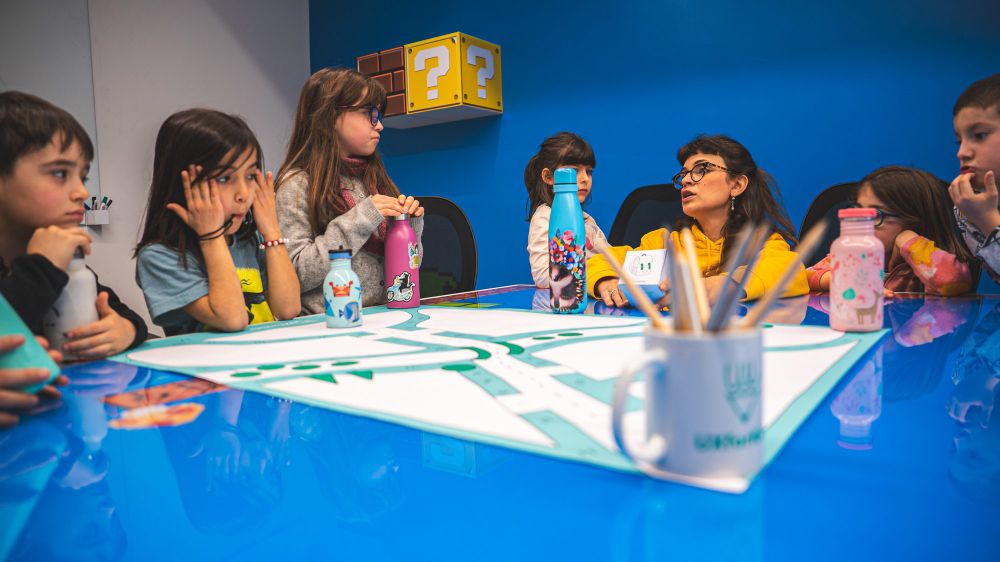 Bambine e bambini attorno al tavolo mentre una mentor mostra la mappa del progetto