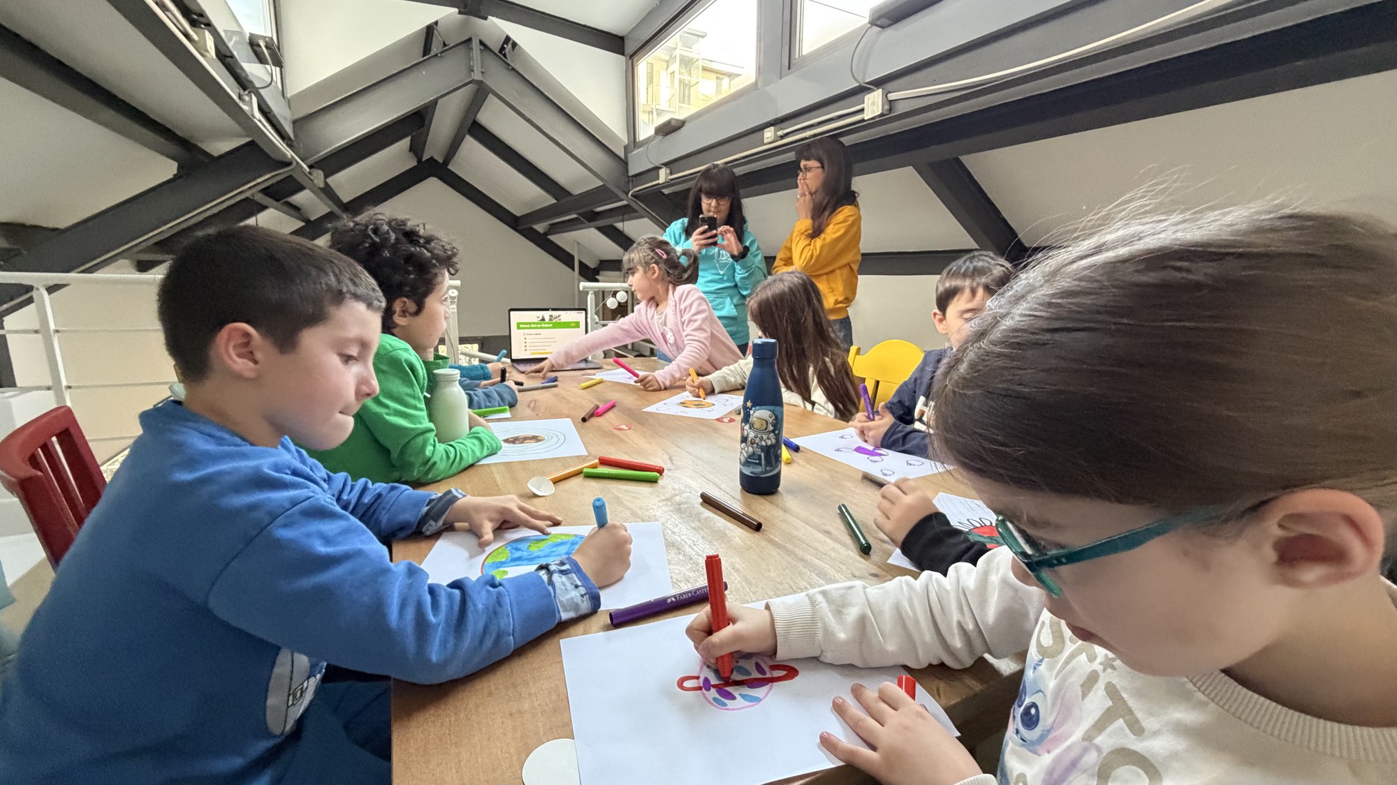 Bambine e bambini disegnano forme e pianeti sui loro fogli, seduti al tavolo lungo