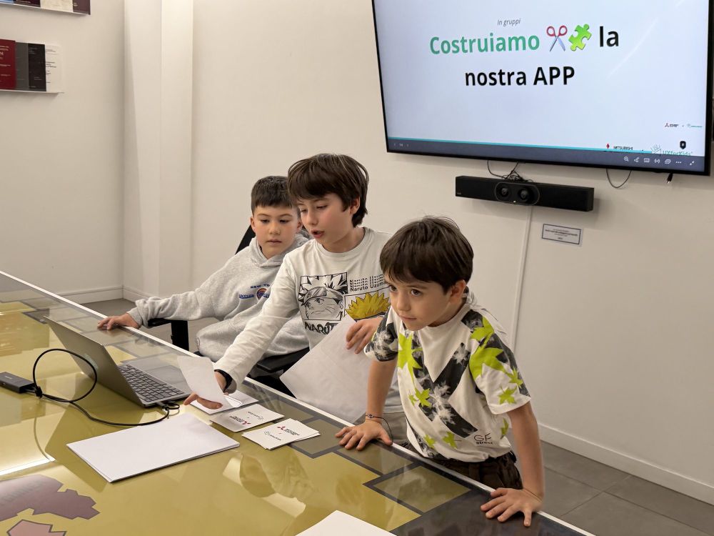 Tre bambini al laptop davanti allo schermo con la scritta “Costruiamo la nostra APP”