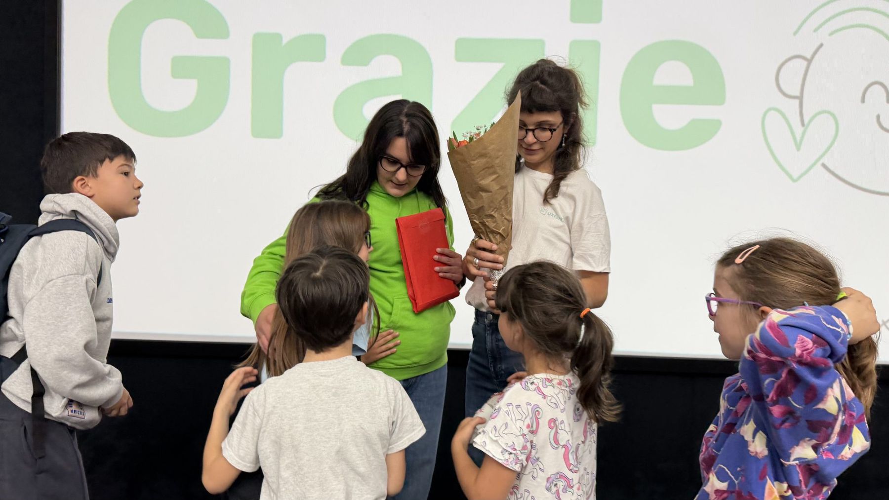 Bambine e bambini consegnano fiori e un regalo alle mentor sul palco, davanti allo schermo con la scritta Grazie